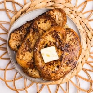 Three slices of french toast stacked on a plate, topped with a pat of butter, and drizzled with maple syrup.