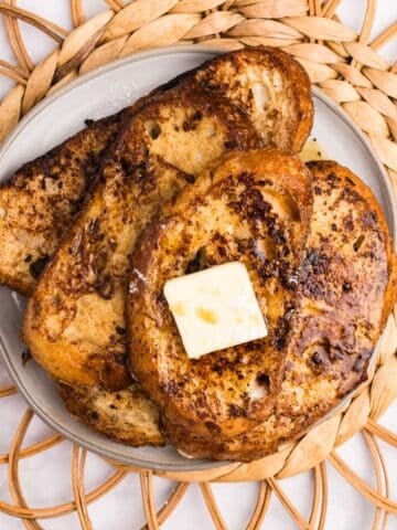 Three slices of french toast stacked on a plate, topped with a pat of butter, and drizzled with maple syrup.